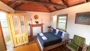 A bedroom with a wooden ceiling, a bed with a striped blanket, a green armchair, a wooden nightstand, and a window with a view of the outdoors.