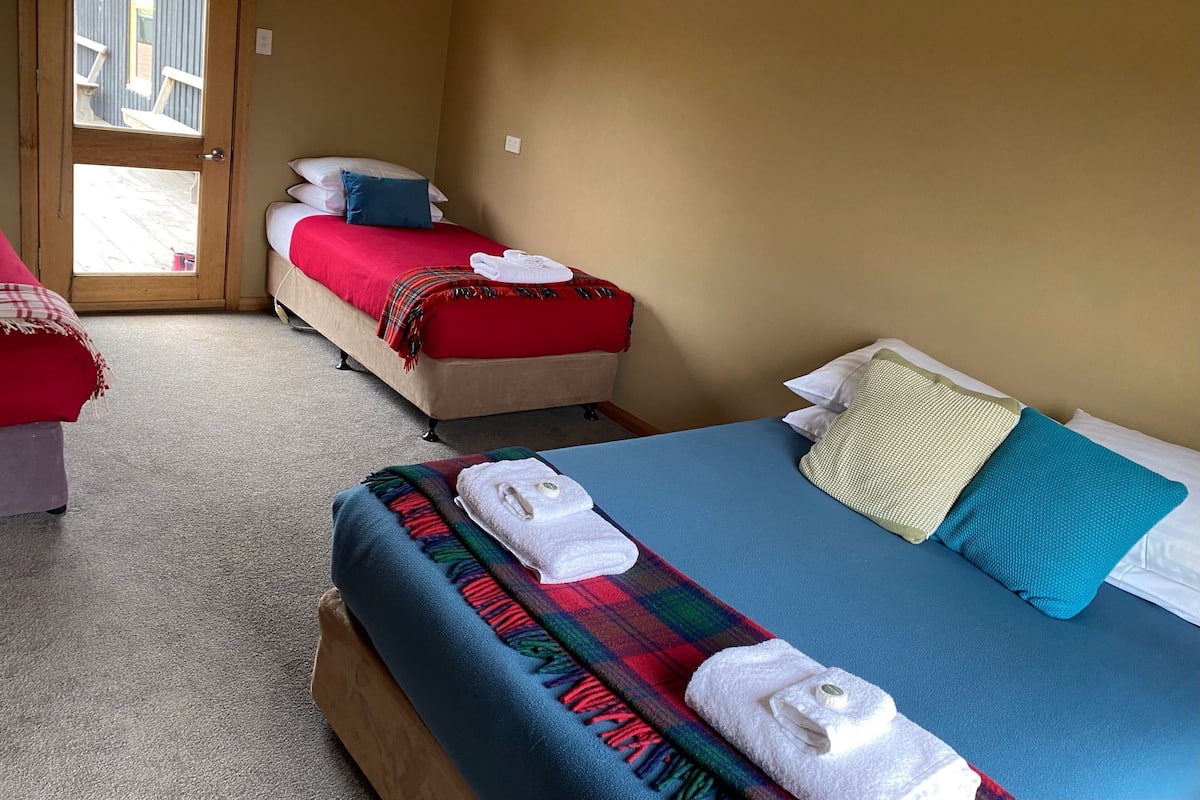A hotel room with two beds, each with a red blanket and pillows, a blue bed with a green pillow, and a wooden bench with towels.