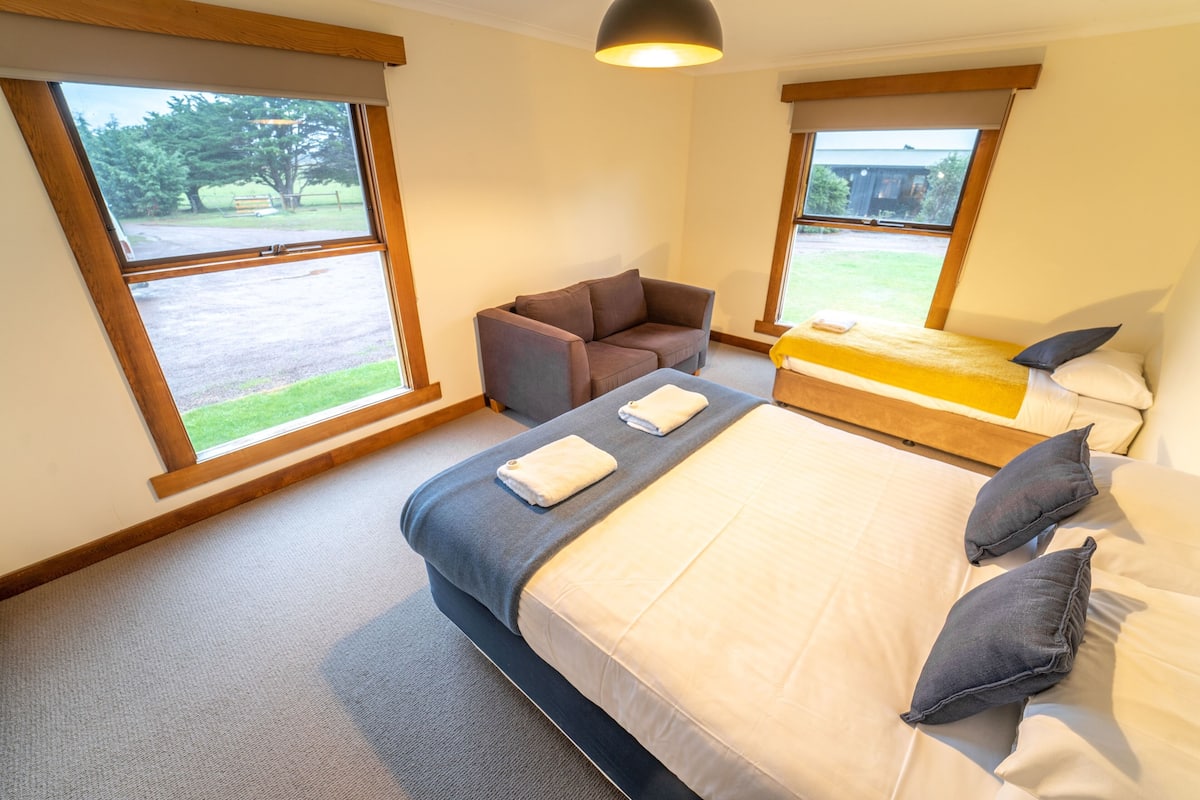 A bedroom with a large bed, a sofa, and two windows with a view of the outdoors.