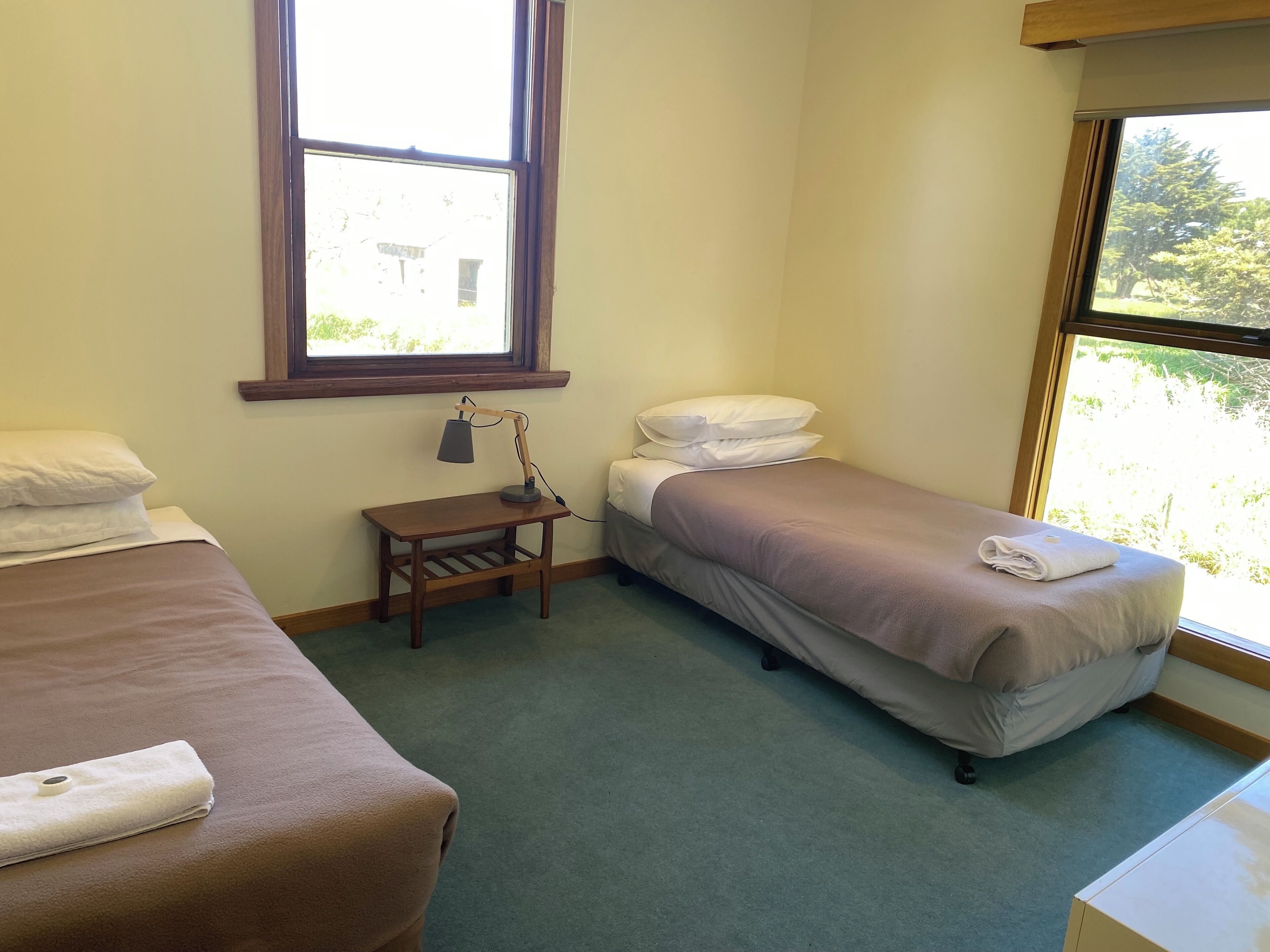 A small, clean room with two single beds, a small table with a lamp, and a window providing a view of greenery outside.