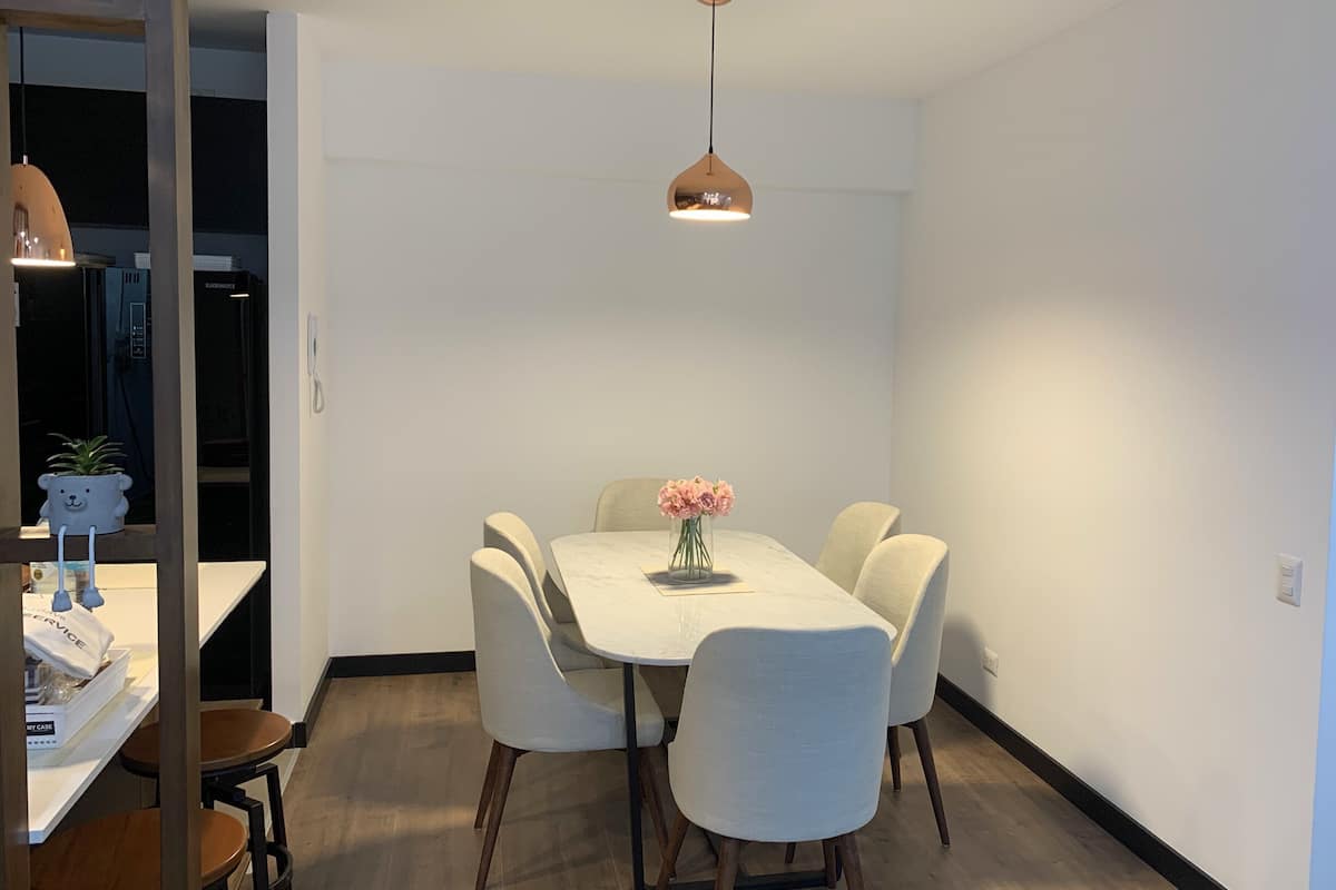A dining area with a white table and chairs, a hanging pendant light, and a vase with pink flowers.