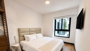 Habitación de hotel con cama, mesita de noche, televisor montado en la pared y ventana con vistas a edificios.