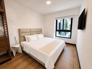 Habitació d'hotel amb llit, taula de nit, televisió penjada a la paret i finestra amb vista a edificis.