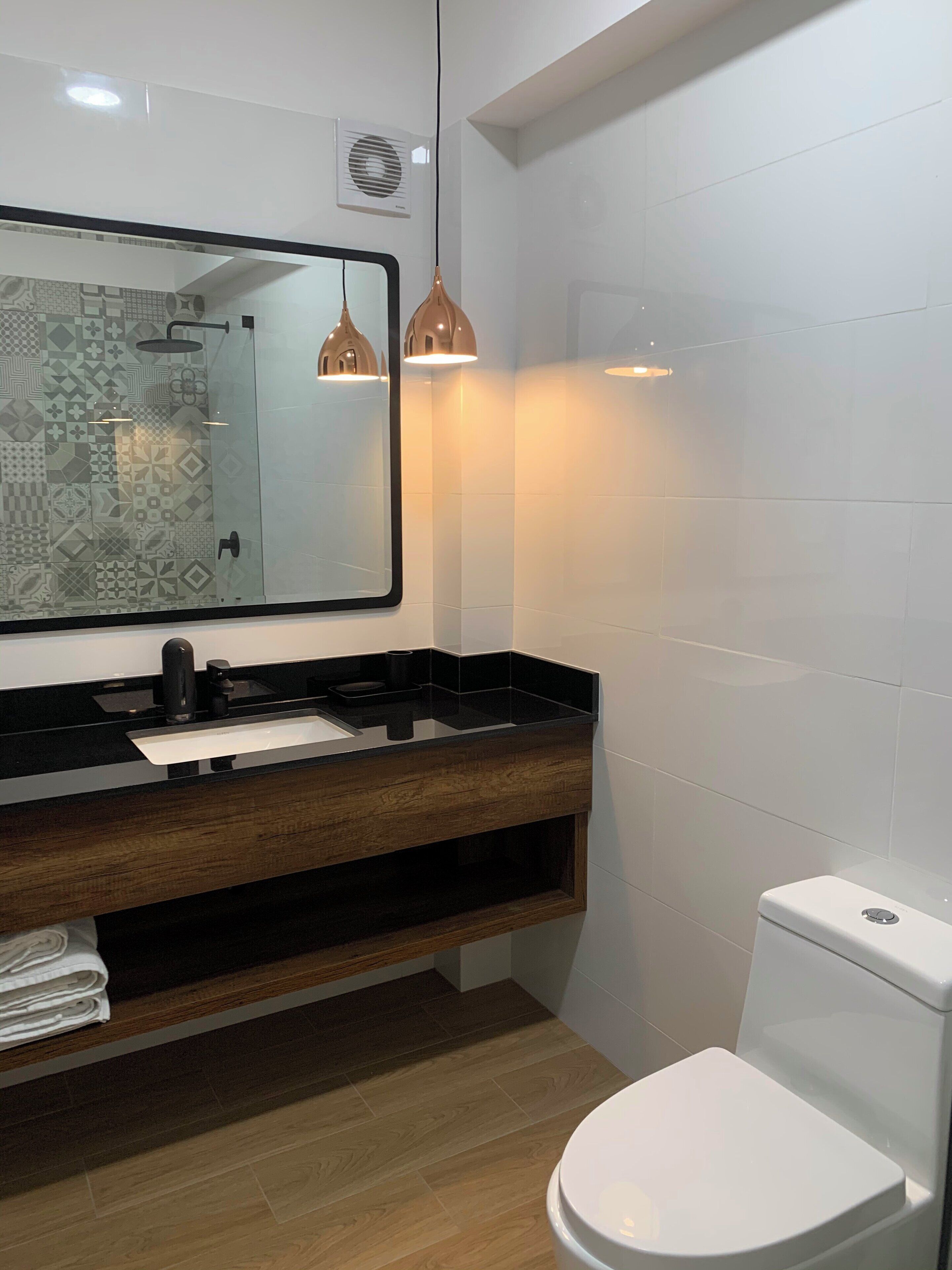 Et moderne badeværelse med et glasbadeværelsesafskærmning, en trækommode med sort bordplade, et hvidt toilet og hængende lysarmaturer.