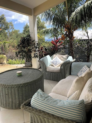 A covered outdoor seating area with wicker furniture, a round table, and a view of tropical vegetation and a building in the background. - Black River Villa Turquoise 3bdr, sea View (Black River)