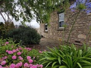 Un edificio de piedra con una ventana y una puerta, rodeado de abundante follaje y plantas en flor.