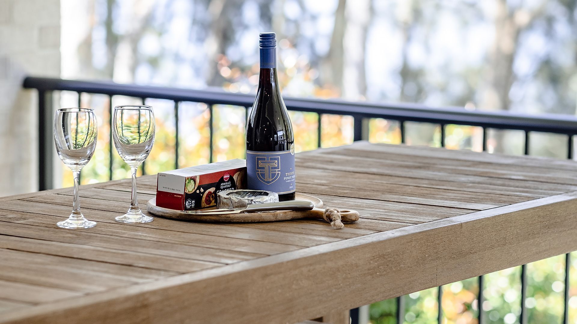 A wooden table with a bottle of wine, two glasses, a box of chocolates, and a corkscrew.