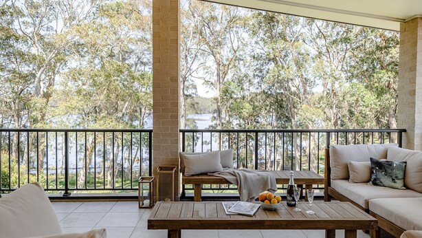 Un balcon avec une table en bois, un canapé et un banc. On aperçoit des arbres et une étendue d'eau au loin.