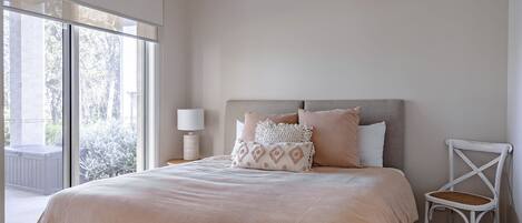 A bedroom with a large window, a bed with a beige bedspread, and a white chair.