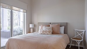 A bedroom with a large window, a bed with a beige bedspread, and a white chair. - Aria Shores - Your Lakeside Haven (Kings Point)