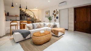 Una sala de estar moderna con un sofá, una mesa de centro y sillas. Se ve una escalera que conduce a una cocina y un comedor.
