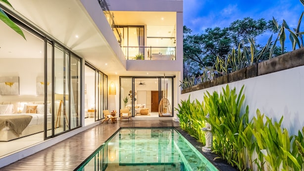 Una casa moderna con piscina de fondo de cristal, un balcón y vistas del entorno verde.