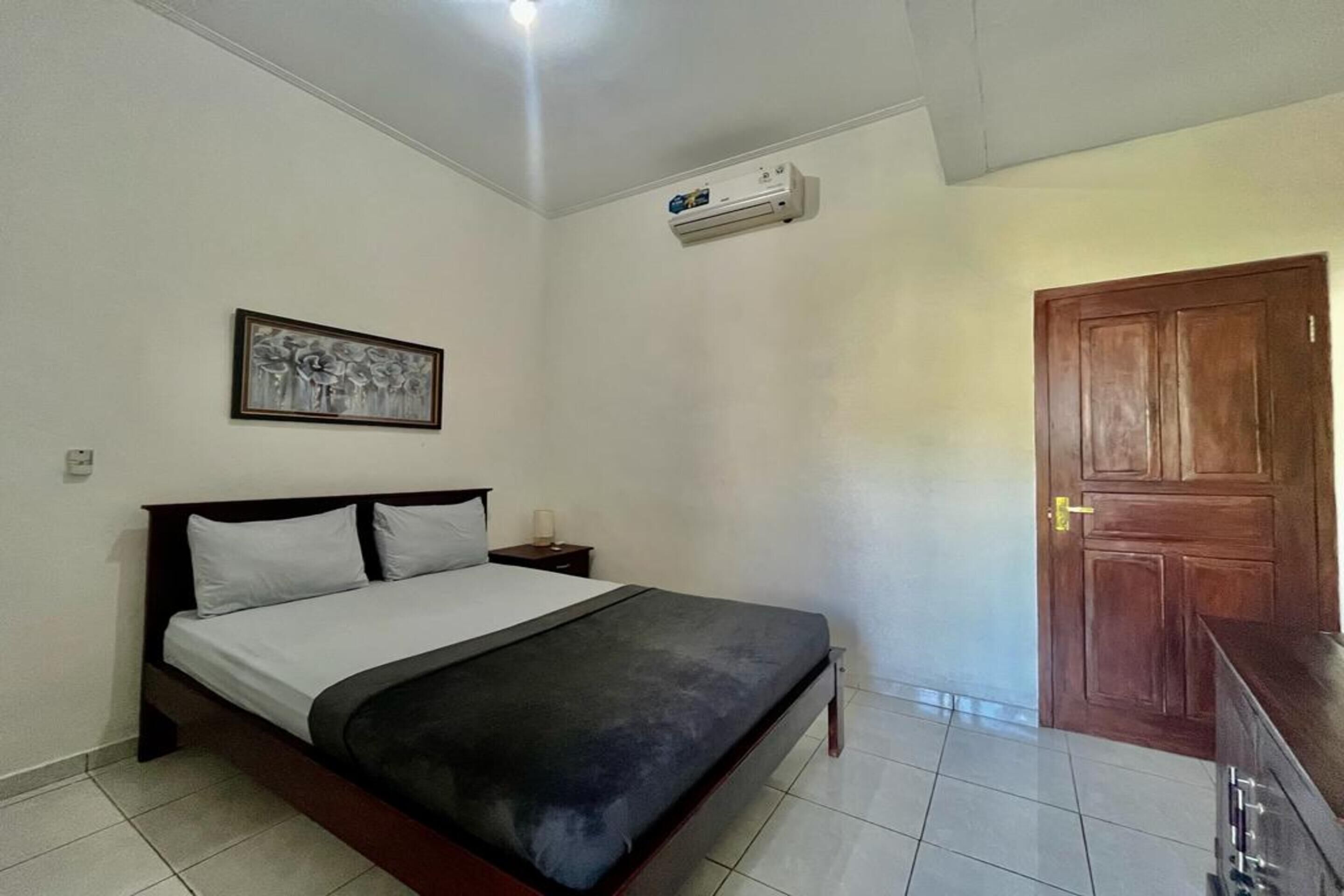 A bedroom with a bed, a nightstand, an air conditioning unit, a wooden door, and a wall-mounted picture.
