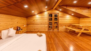 A cozy wooden cabin room with a bed, a table, and a bench. - The Oaknut Cottage Birding Lodge (Kosiyakutoli)
