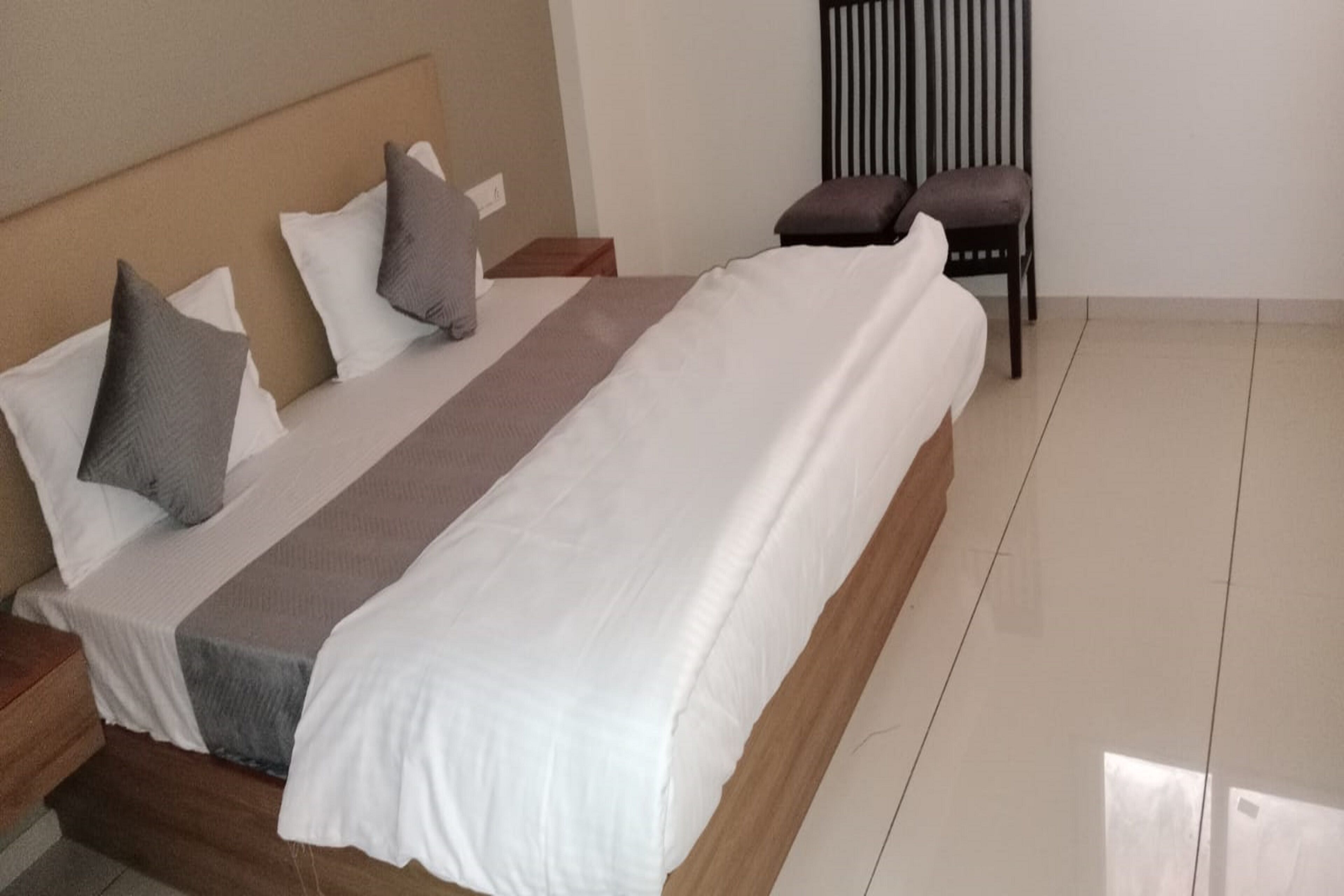 A neatly made bed with a white and gray bedspread, two pillows, and a wooden headboard. There is a wooden nightstand with a lamp and a small table beside the bed. A chair is positioned against the wall.