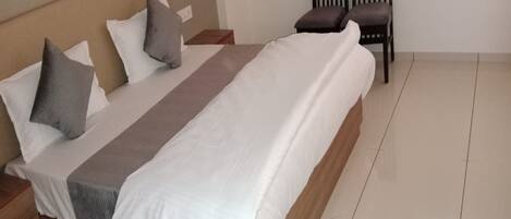 A neatly made bed with a white and gray bedspread, two pillows, and a wooden headboard. There is a wooden nightstand with a lamp and a small table beside the bed. A chair is positioned against the wall.