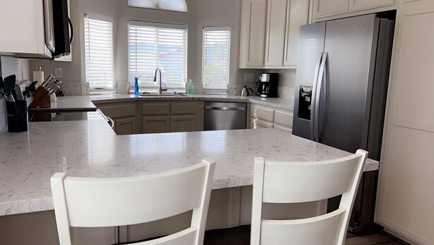 A kitchen with a large island, white cabinets, a stainless steel refrigerator, and a window with blinds.