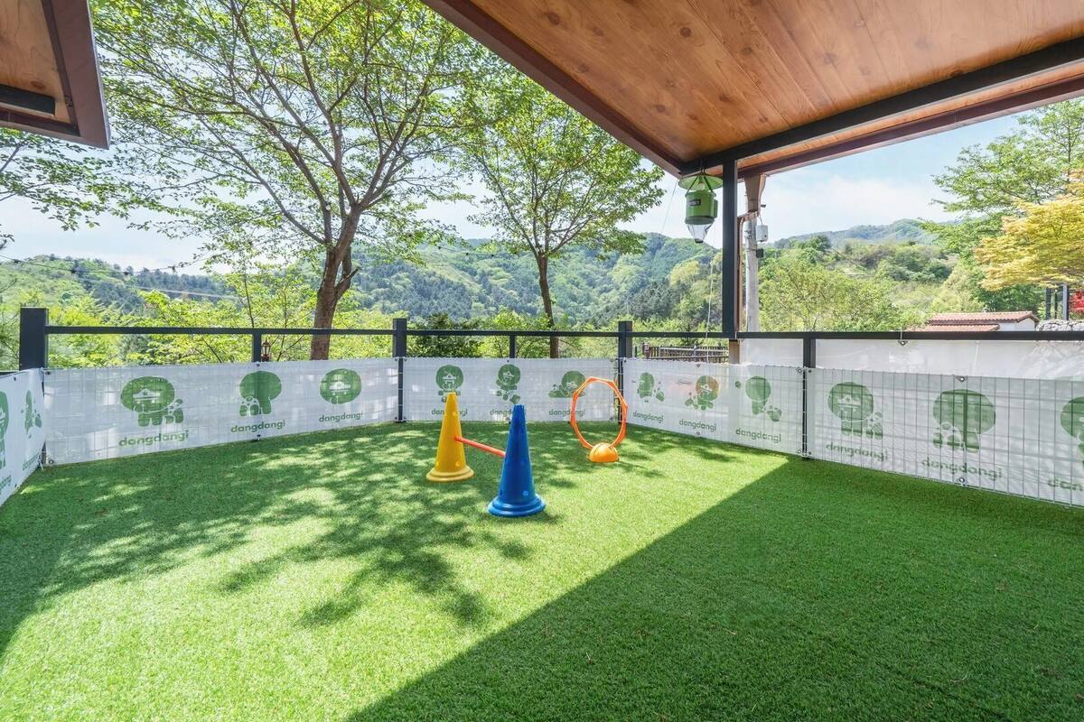 Un espace extérieur doté d’une pelouse artificielle, d’une pergola en bois et offrant une vue sur un paysage boisé.