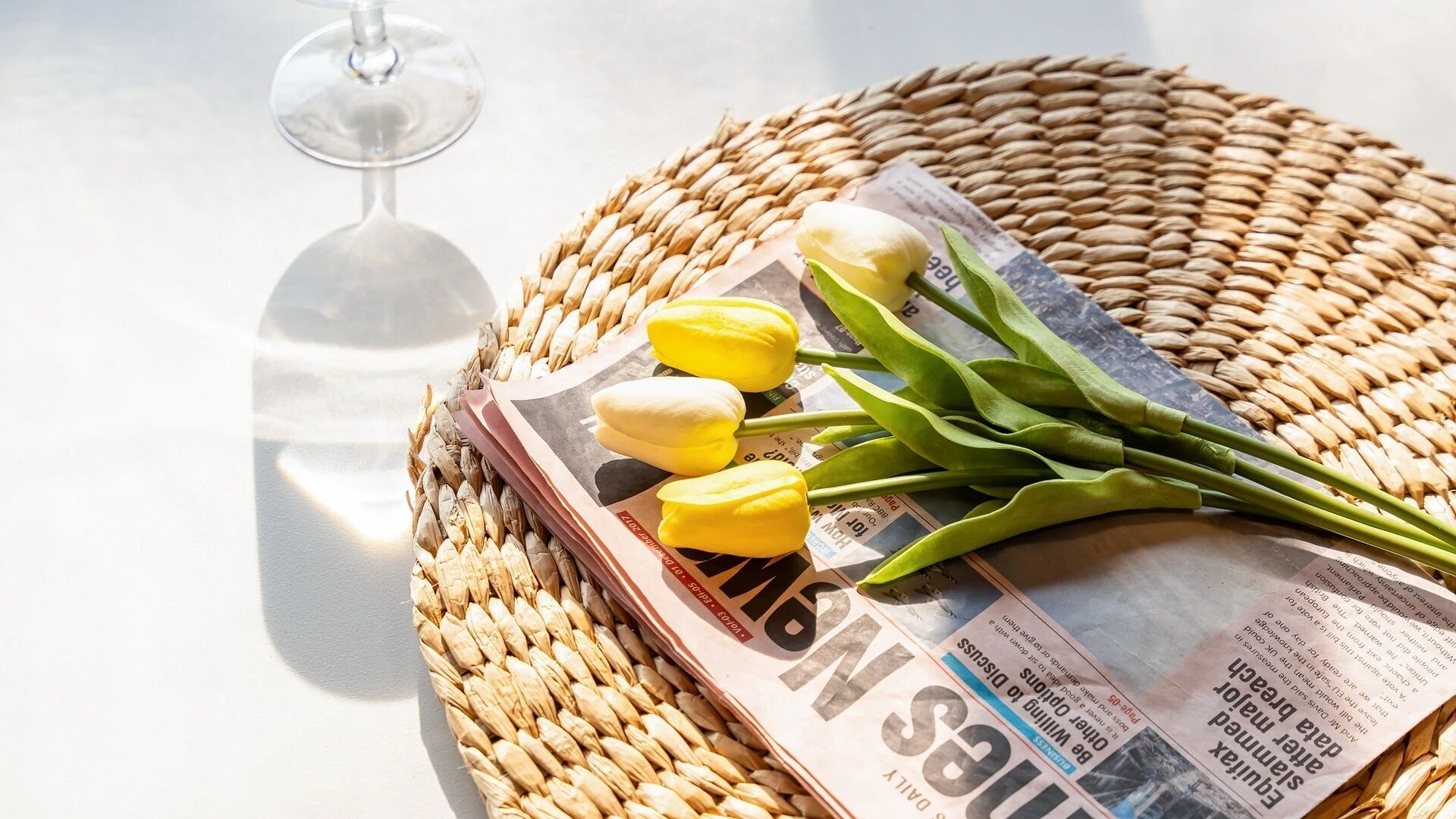 A woven mat, a glass of wine, and yellow tulips on a table.