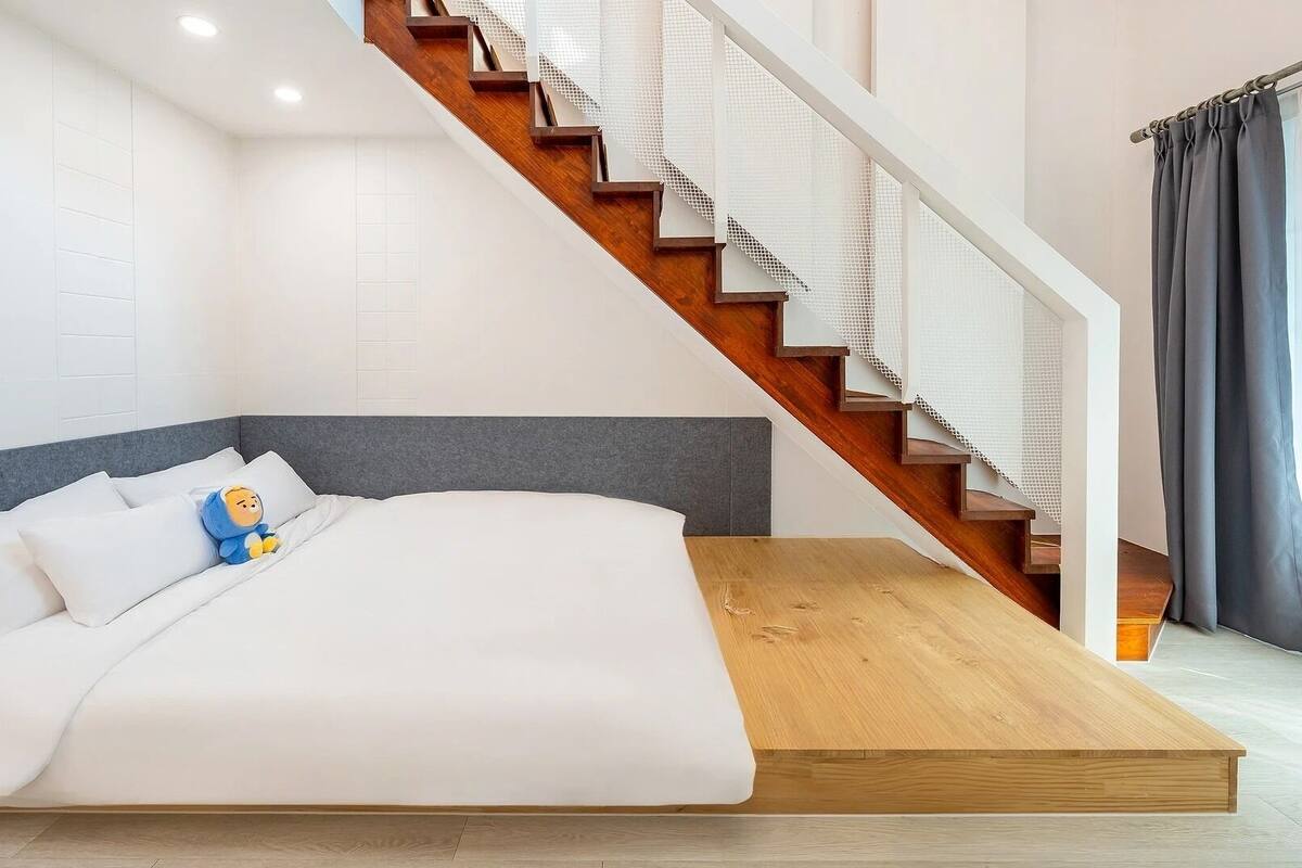 Une chambre moderne avec un lit, une tête de lit en bois, un escalier avec une rampe blanche et une fenêtre avec des rideaux.