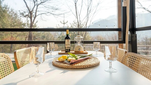 Une table dressée avec des verres à vin, une bouteille et une bougie, offrant une vue sur les arbres et les montagnes à travers de grandes fenêtres.