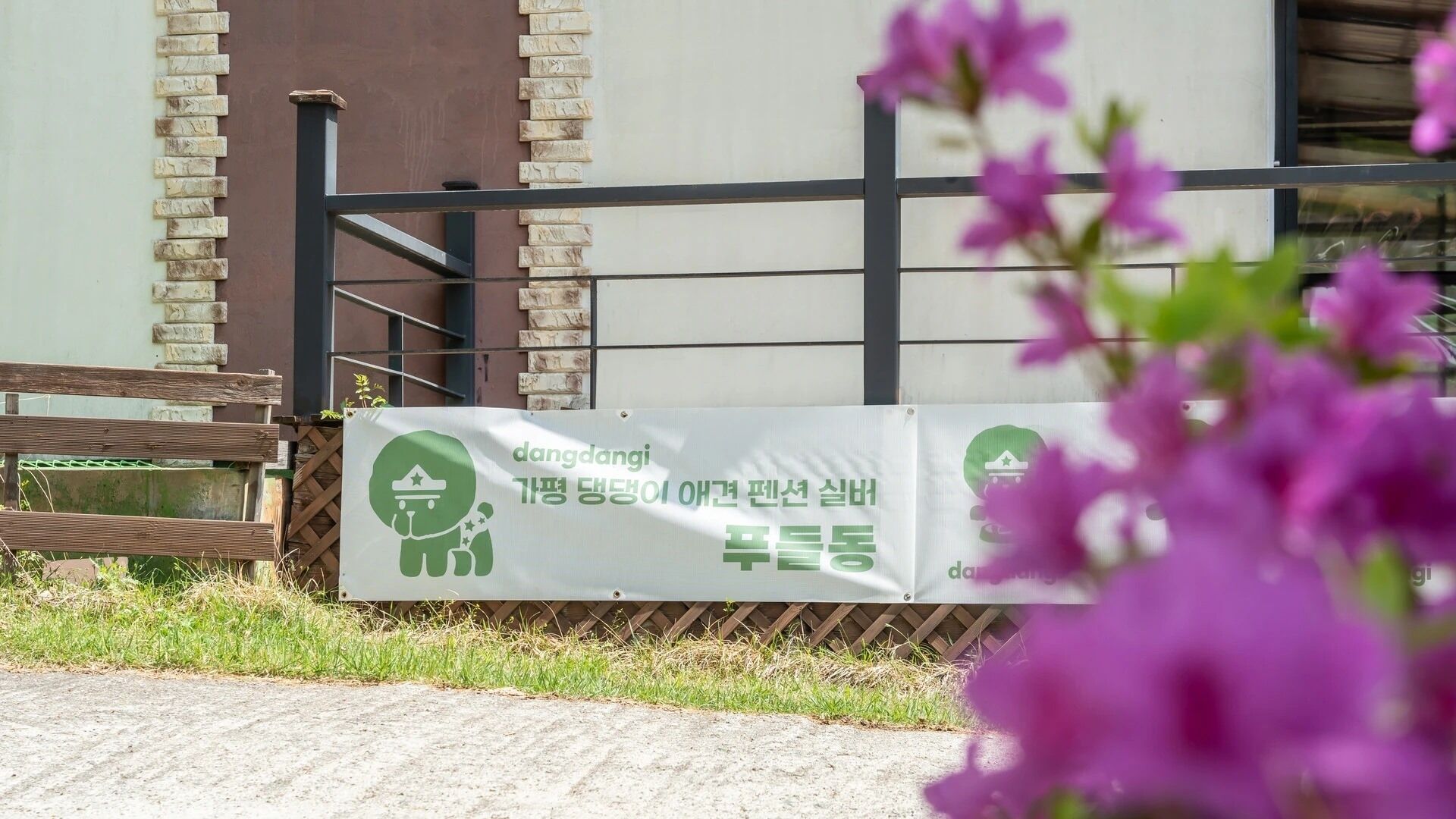 A banner with Korean text and a logo, a wooden bench, and a building with a brick wall and metal railing.