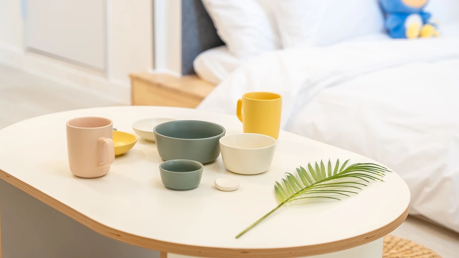 A neatly arranged table with various ceramic mugs and bowls, a single green leaf, and a bed in the background.