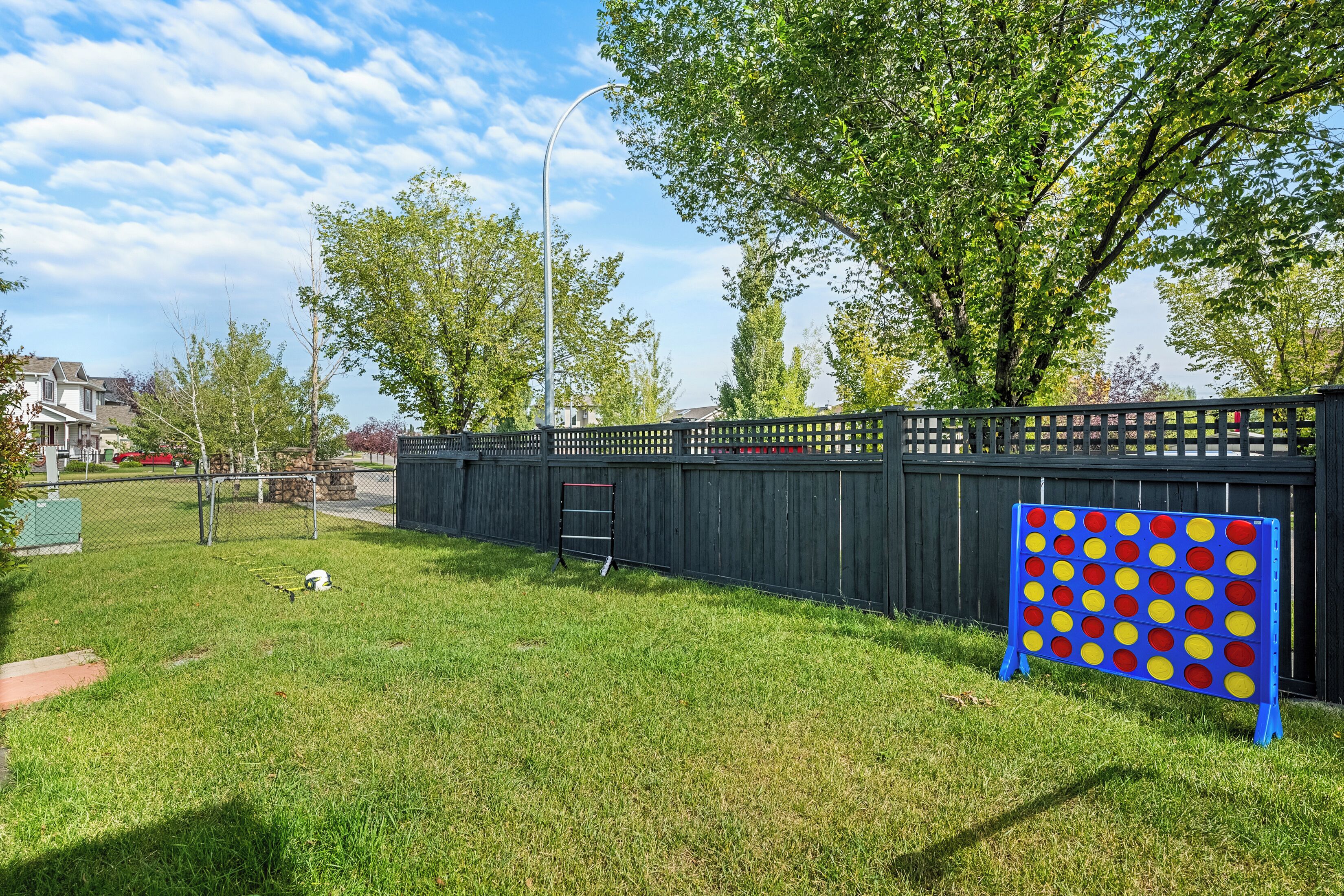 A fenced backyard with a blue and yellow board game, a ladder, and a small white object on the grass.