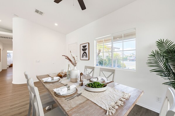 A dining area with a wooden table set for four, white chairs, a large window with a view, and a potted plant on the right.
