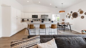 A spacious kitchen with white cabinets, a refrigerator, and a microwave. There are three chairs and a dining table with chairs. The room has a large window and a hanging light fixture.