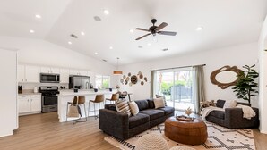 A modern kitchen with white cabinets, a central island, and a dining area with chairs. There is a dark-colored sofa and ottoman in the living area, and a large window providing natural light.