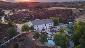 A large white building with a grey roof, surrounded by a fenced area with a pool and a tennis court. - Expansive Wine Country Estate | Pool + Hot Tub | Sleeps 20 | Quiet Retreat (Temecula)