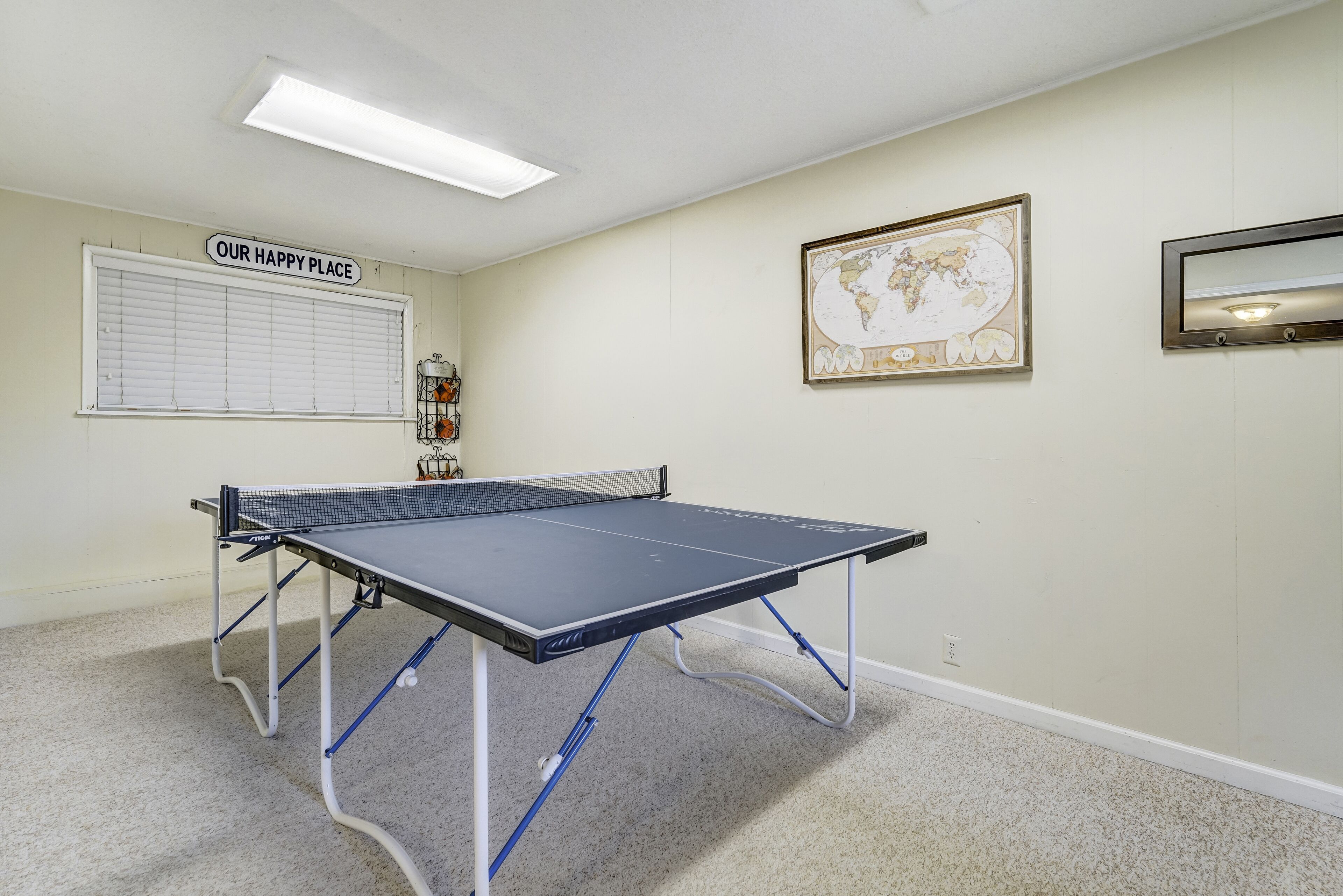 A ping pong table in a room with a world map on the wall, a window with blinds, and a sign reading "OUR HAPPY PLACE".