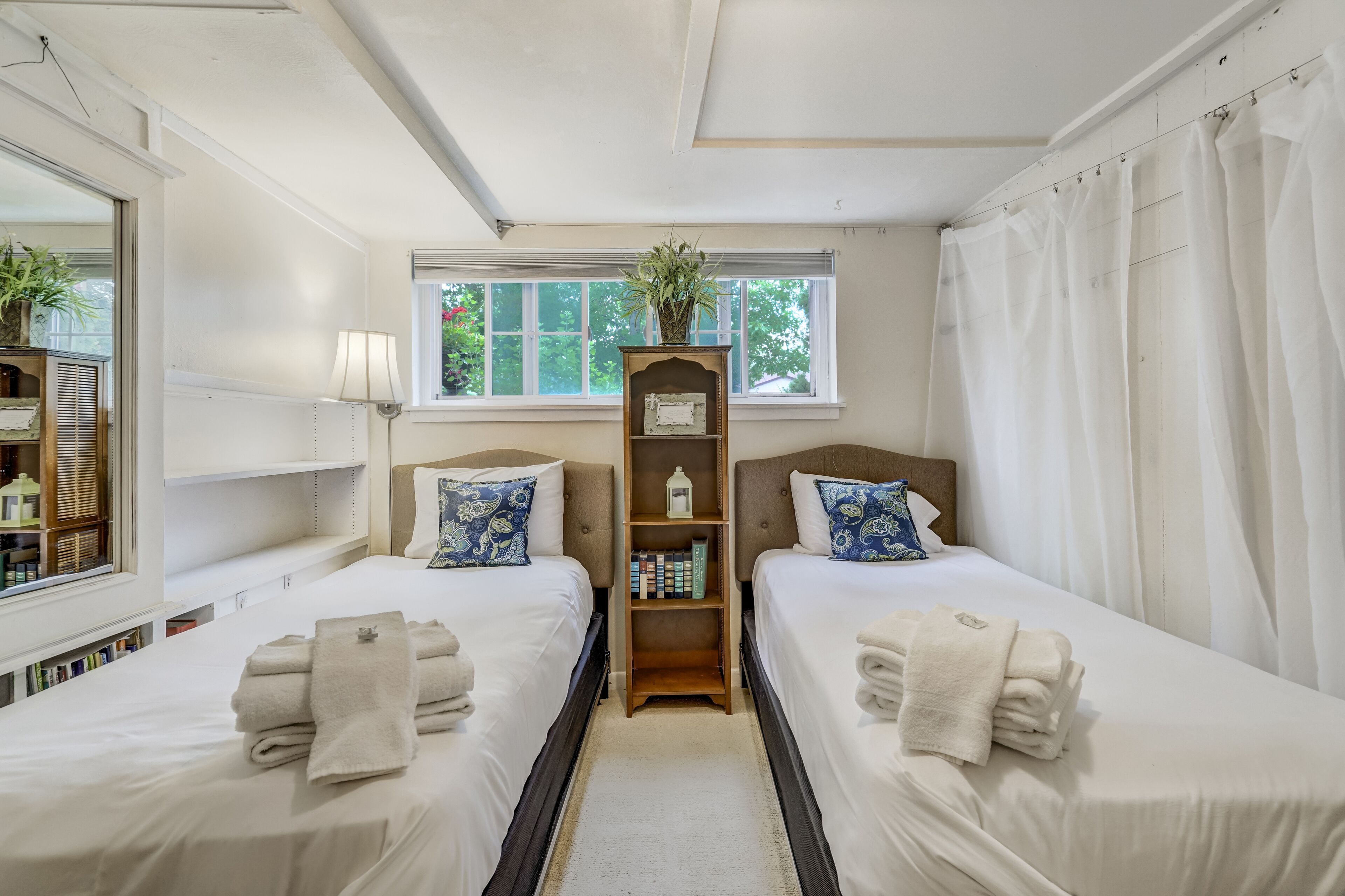 A hotel room with two beds, a window with a view, a bookshelf, and a plant.