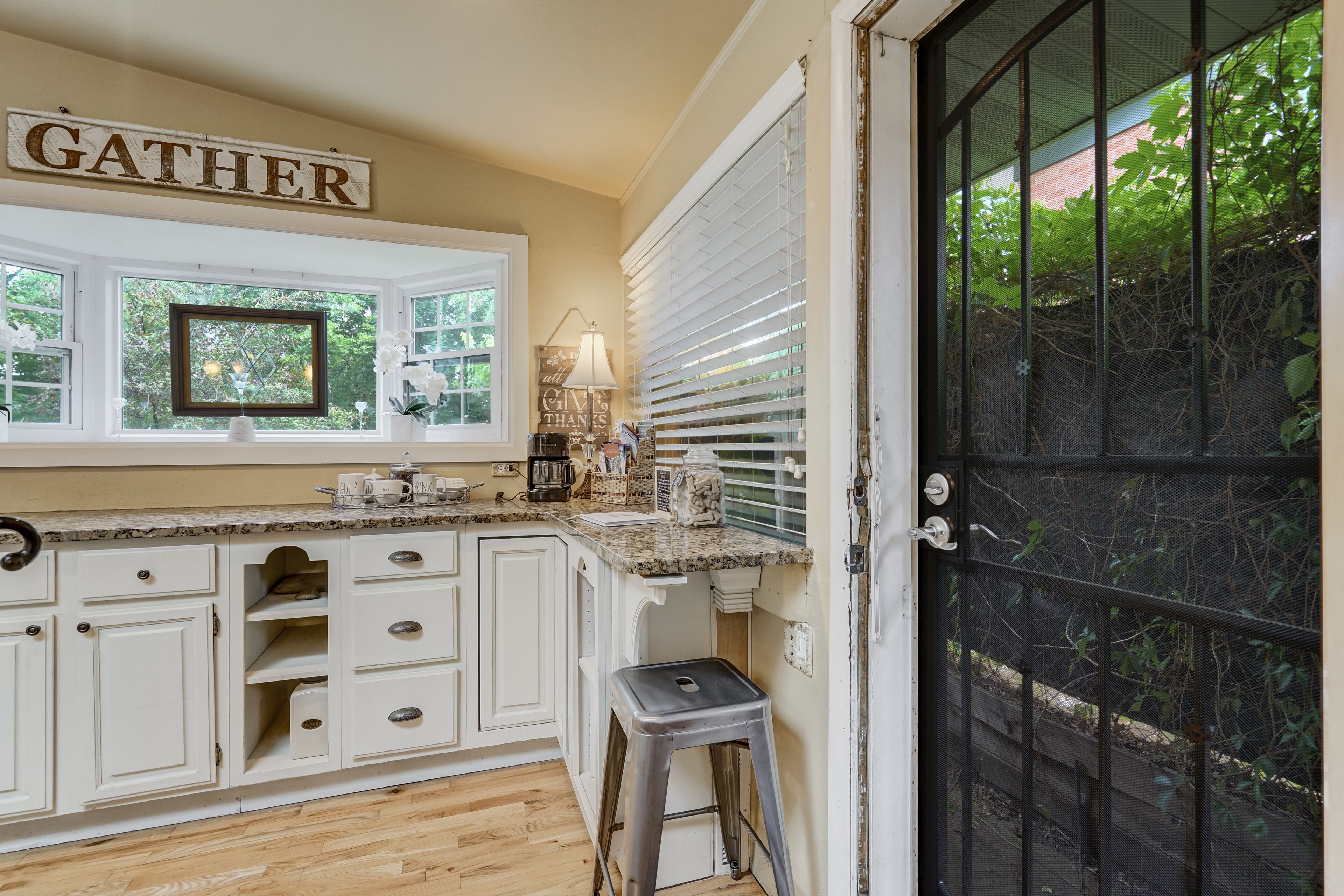 Una cocina con armarios blancos, una ventana con vistas, un letrero que dice 'GATHER' y una puerta con mosquitero.