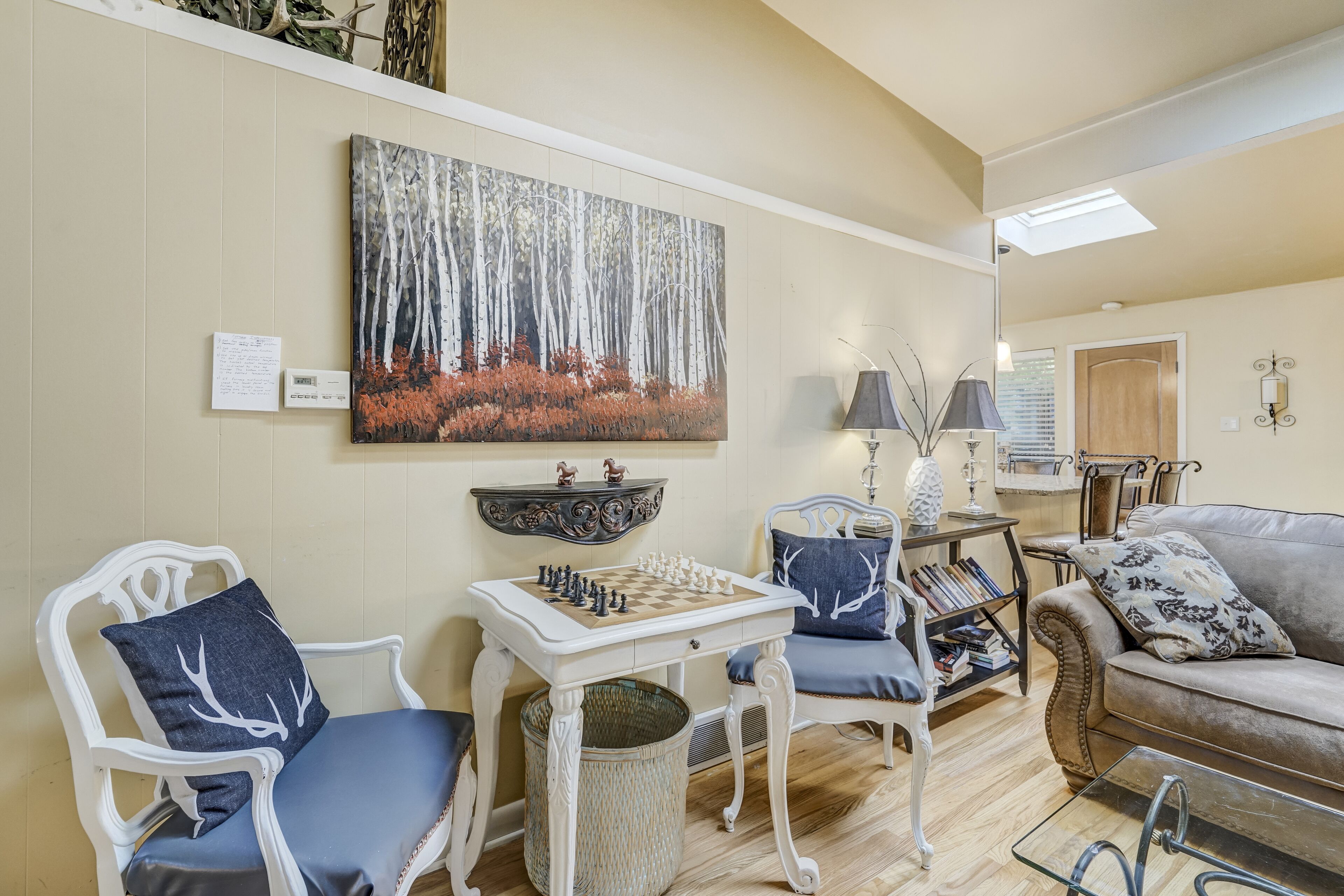 A cozy living room with a chessboard, two chairs, a sofa, a bookshelf, and a painting of a forest.
