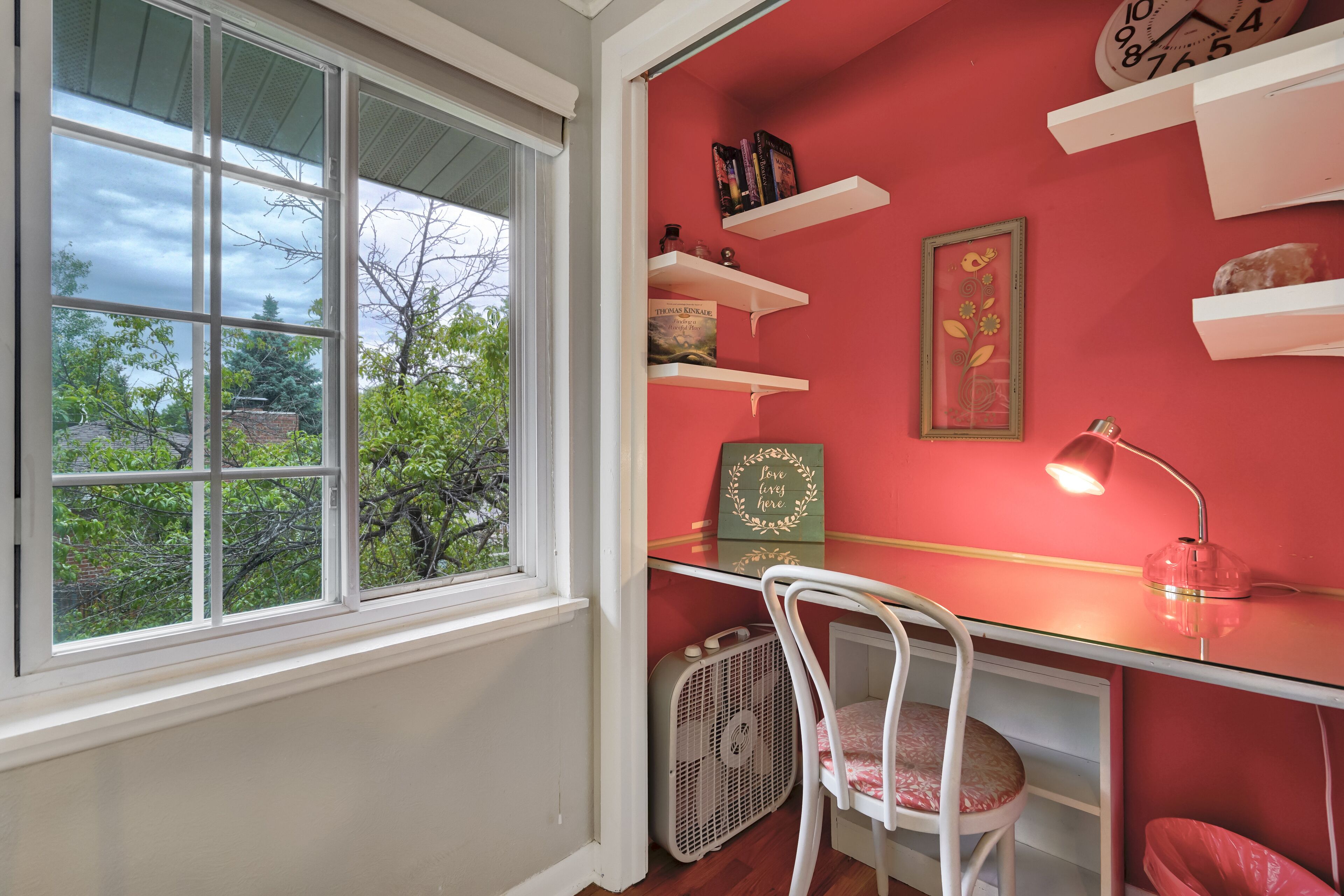 A room with a desk, a chair, a fan, and shelves with books and decorations.