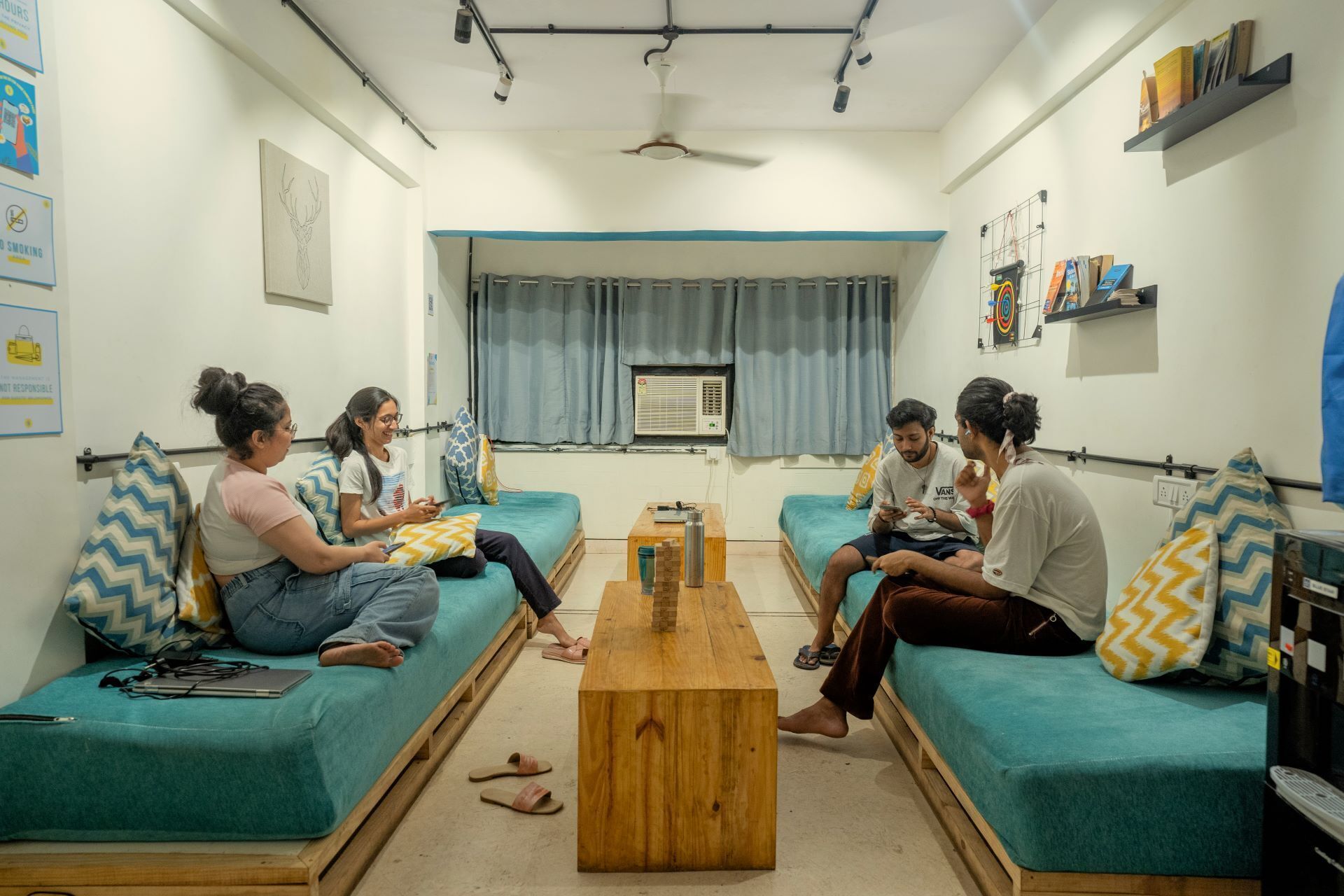 Ein Zimmer mit zwei Sofas, einem Couchtisch und einem Deckenventilator. An den Wänden hängen Poster und es gibt eine Klimaanlage.