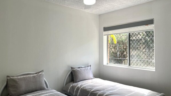 Un dormitorio con dos camas individuales, una ventana con mosquitero y una pared blanca lisa.