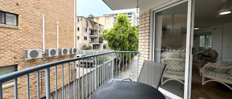 Een balkon met een metalen hekwerk, een zwarte tafel en een schuifdeur die leidt naar een ruimte met rieten meubels.