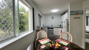 A dining area with a table set for two, a kitchen in the background, and a view through a window. - Chequers Caloundra U2, Walk To Kings & Shelly (KINGS BEACH)
