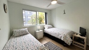 Un dormitorio con dos camas, una ventana, un ventilador de techo, un televisor y una pequeña mesa.