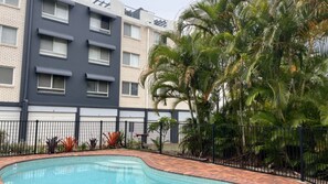 Una piscina con borde de ladrillo, rodeada por una valla, contigua a un edificio de varios pisos con balcones.