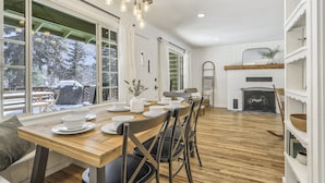 A dining area with a wooden table and chairs, a fireplace, and large windows with a view of trees. - Green Dream (Cascade)