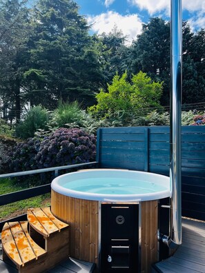 Een hottub met houten zitplaatsen en een metalen paal, omgeven door weelderige begroeiing en een houten terras.