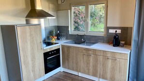 A compact kitchen with wooden cabinets, a built-in oven, a sink, and a window overlooking greenery. - Chalet - Terrasse & bain nordique (Trégastel)