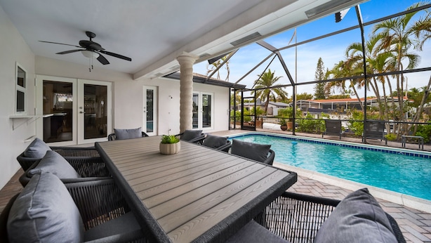 Ein Poolbereich mit Holztisch und Korbstühlen, einem Deckenventilator und Blick auf den Pool und die umliegenden Palmen.