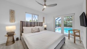 Ein Schlafzimmer mit einem Bett, einem Deckenventilator, einem Nachttisch mit Lampe, einem Holzstuhl und Blick auf einen Swimmingpool durch eine Glastür.