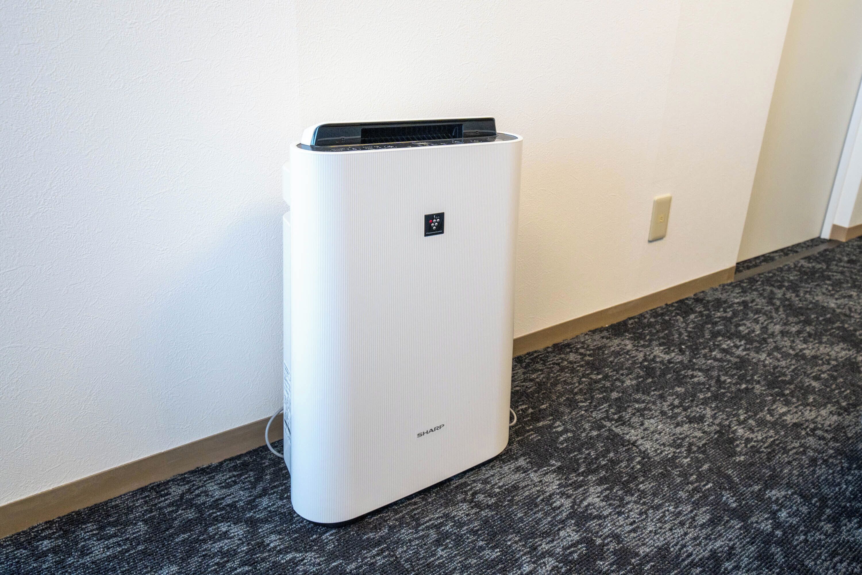 A white air conditioning unit with a control panel on top, placed against a wall in a hallway with a carpeted floor.