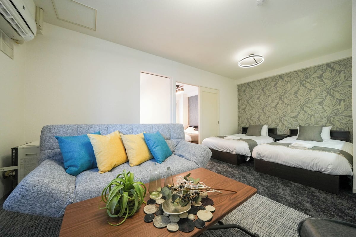 Quarto de hotel com um sofá cinzento, duas camas de solteiro, uma mesa de centro de madeira com objetos decorativos e papel de parede estampado nas paredes.